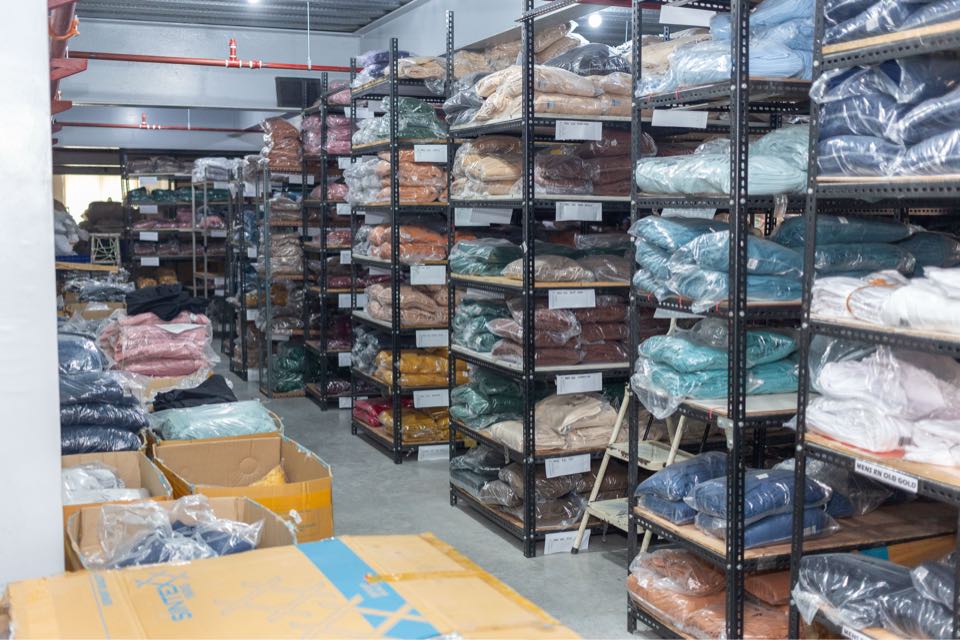 Rows of steel shelving filled with folded finished garments, sorted by color, above cartons on the floor.