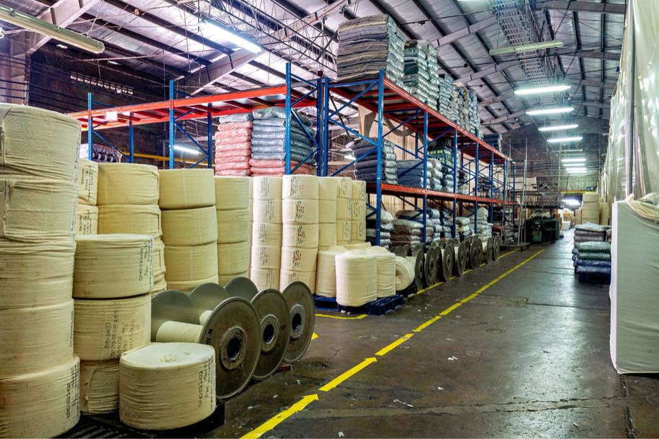 Rolls of undyed greige knitted fabric stacked in front of tall warehouse racks of wrapped fabric.