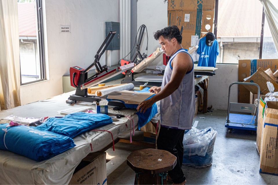 A worker at a heat press station presses a print onto a shirt, with folded blue shirts waiting on the table.